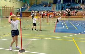  dzieci znajdujące sie na sali gimanstycznej, konkurują w przekładaniu szarfy przez ciało a potem biegu w prawą stronę zdjęcia 