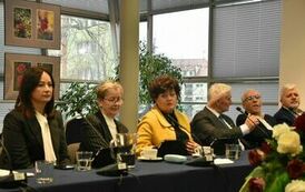 Na środku przy stole siedzi pani w pomarańczowo-brązowych włosach oraz ż&oacute;łtym żakiecie, po jej prawicy siedzą na czarno ubrani panowie z białymi włosami a po lewej na czarno ubrane panie 