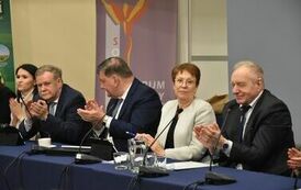 przy stole klaszcze trzech pan&oacute;w w marynarkach oraz pomiędzy nimi pani w białym żakiecie 