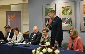 część członk&oacute;w od strony stolik&oacute;w z odą, po środku zgromadzonych obok pani w zielonej koszuli z czarnymi paskami oraz czarnej sukni stoi z mikrofonem pan w cały na granatowo 