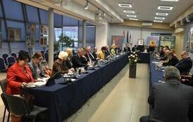 członkowie rady zebrani razem przy stole, po prawej z filiżankami na kawę oraz łakociami, a po prawej panowie i panie ze szklankami wody