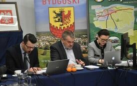 trzy osoby, dwoje mężczyzn i pani przy laptopie appla siedzą przy stole na tle baner&oacute;w powiatu bydgoskiego oraz innych miejscowości 