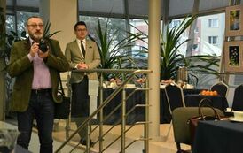 na zdjęciu pośr&oacute;d stolik&oacute;w pan, ky&oacute;ty wcześniej przemawiał w jasnej marynarce, a także bliżej drugi fotograf idący z aparatem