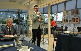 po środku przemawia pan w jasnym garniturze oraz z okularami i mikrofonem, po jego prawej siedzi przy końcu stołu niezadowolony starszy pan w granatowym garniturze a po prawej stronie zdjęcia, w tle znajduje się odwr&oacute;cona plecami pani z czerwonym kubraczk