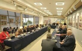 z kadry od strony rogu pokoju, członkowie rady zebrani razem przy stole, po prawej z filiżankami na kawę oraz łakociami, a po prawej panowie i panie z dokumentami