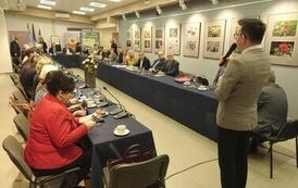 członkowie rady zebrani razem przy stole, po prawej z filiżankami na kawę oraz łakociami, a po prawej panowie i panie z dokumentami. Wszyscy patrzą w stronę przemawiającego  przed stołem pana z miktrofonem