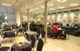 Kilka rozłożonych stolik&oacute;w z jedzeniem oraz krzesłami a za nimi członkowie rady zebrani razem przy stole, po prawej z filiżankami na kawę oraz łakociami, a po prawej panowie i panie z dokumentami.