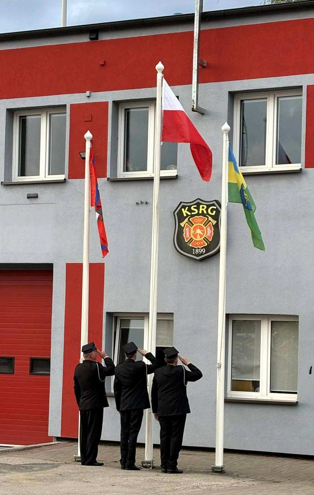 trzech_strażak&oacute;w_salutujących_przed_flagami